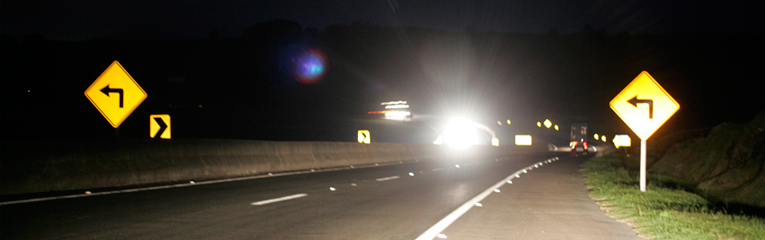 Estrada durante a noite, faróis no horizonte ofuscam a visão.