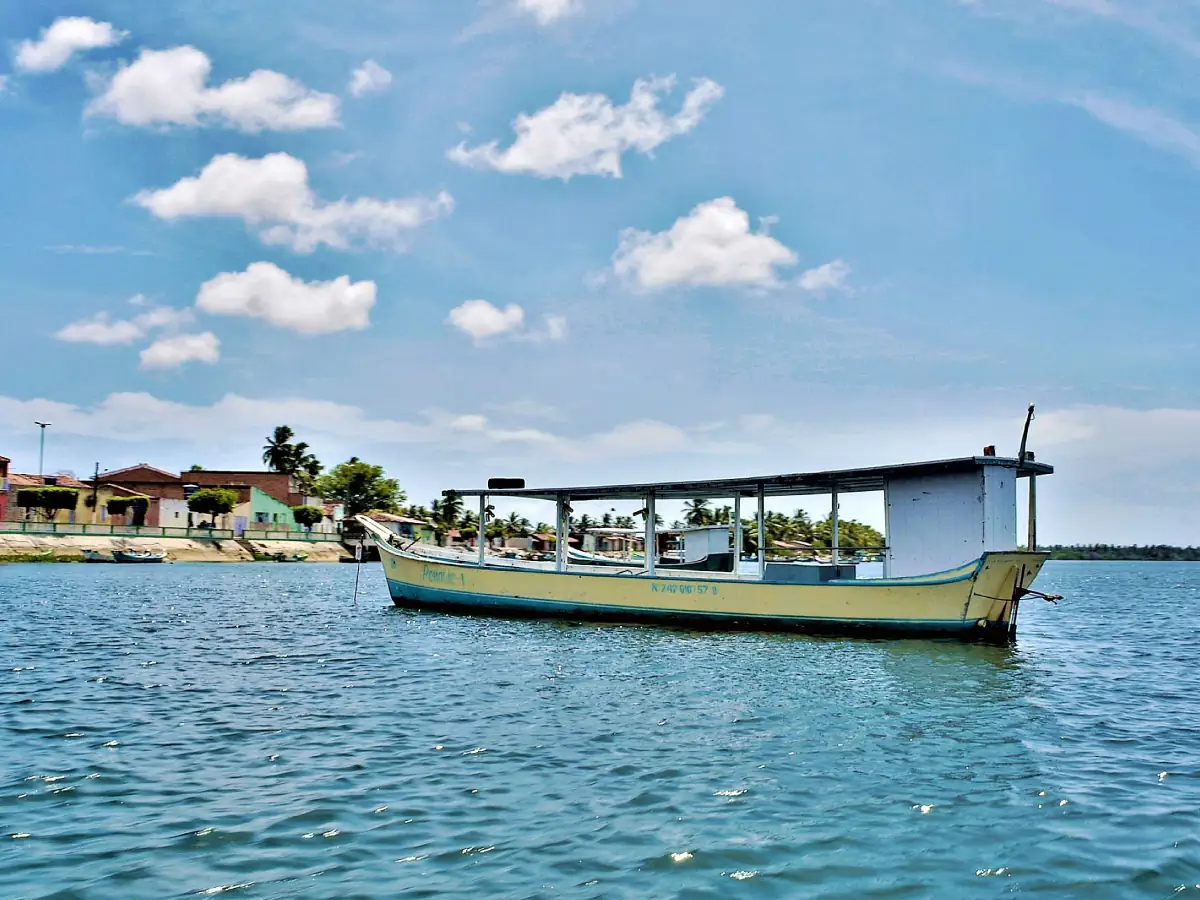 Barco segue ancorado no Rio São Francisco. Ao fundo, há casas pertencentes à cidade de Piaçabuçu. O céu está límpido, com algumas formações de nuvens.