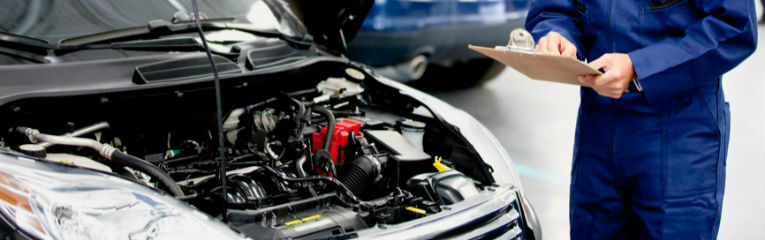 Um homem segura uma prancheta ao lado de um carro com o capô aberto, expondo o motor do veículo.