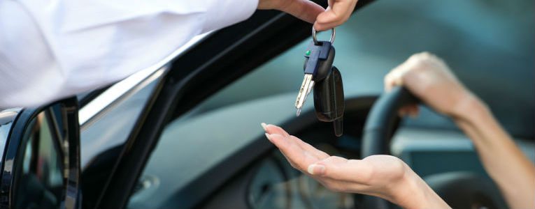 Uma mão entrega as chaves de um carro para uma pessoa sentada no banco do motorista de um veículo.