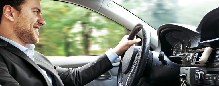 Homem sorri enquanto dirige um carro. Ao fundo, é possível ver árvores desfocadas pela janela.
