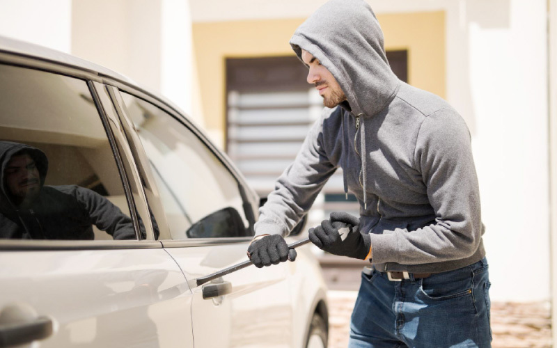 Suposto criminoso com a cabeça envolta por toca, utilizando um artefato para tentar abrir uma das portas de um carro.