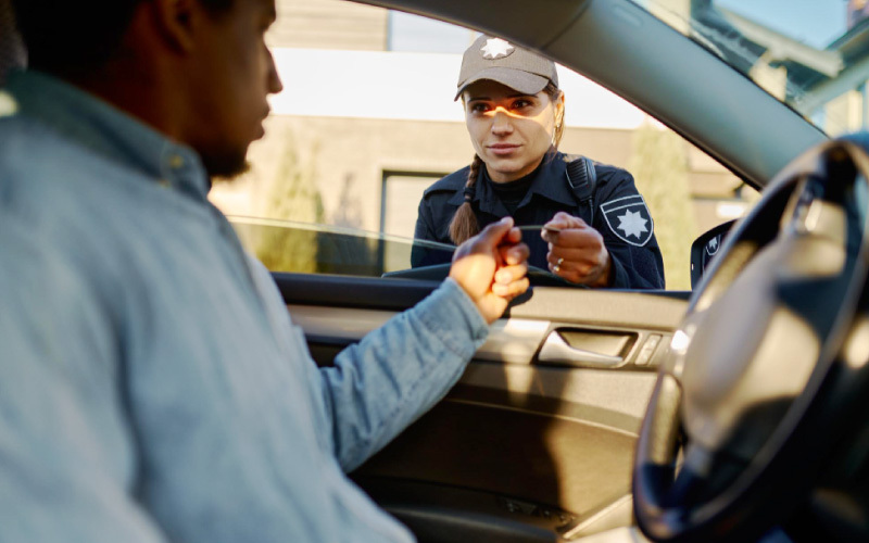 Homem no interior de um veículo recebendo documento de policial feminina que segue do lado de fora.