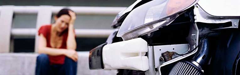 Frente de um carro amassada, uma mulher está sentada com uma expressão preocupada ao fundo.