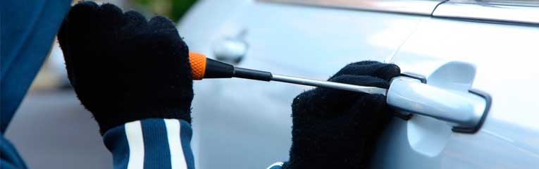 Um homem usando luvas pretas tenta arrombar a porta de um carro usando uma chave de fenda.
