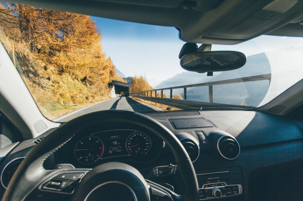A imagem mostra a vista da janela frontal de um carro. É possível ver o painel do veículo e a estrada à frente, com árvores outonais nas laterais e o céu azul ao fundo.