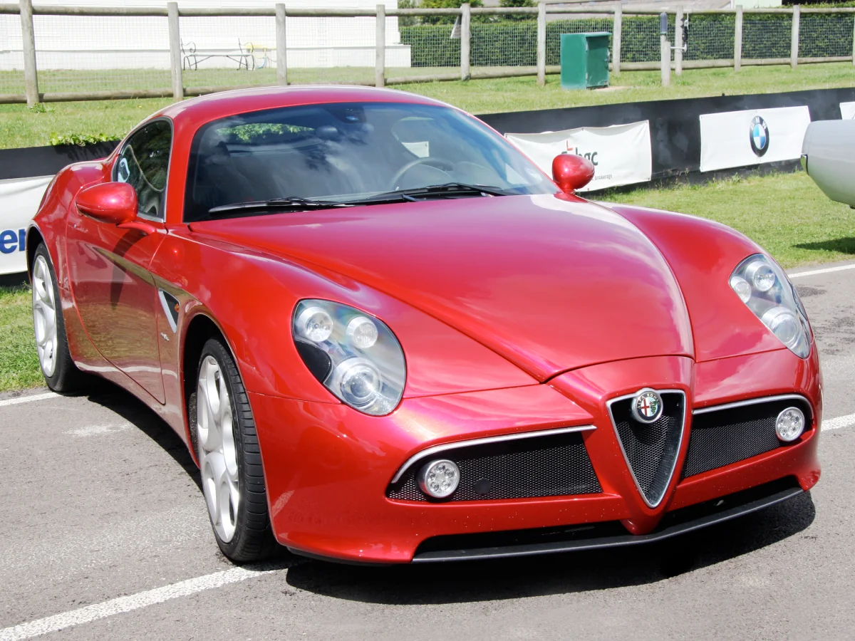 Alfa Romeo 8C Competizione (2007) na cor vermelha, estacionado em área externa sobre uma pista, com cercado de madeira e banners ao fundo.