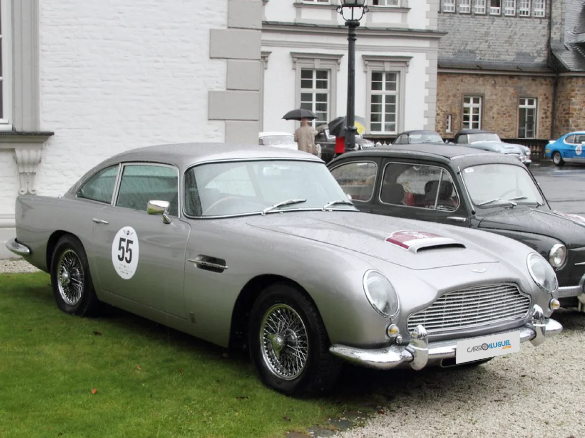 Aston Martin DB5 (1963 - 1965) na cor prata, estacionado sobre gramado, com prédio antigo e pessoas sob guarda-chuvas ao fundo.