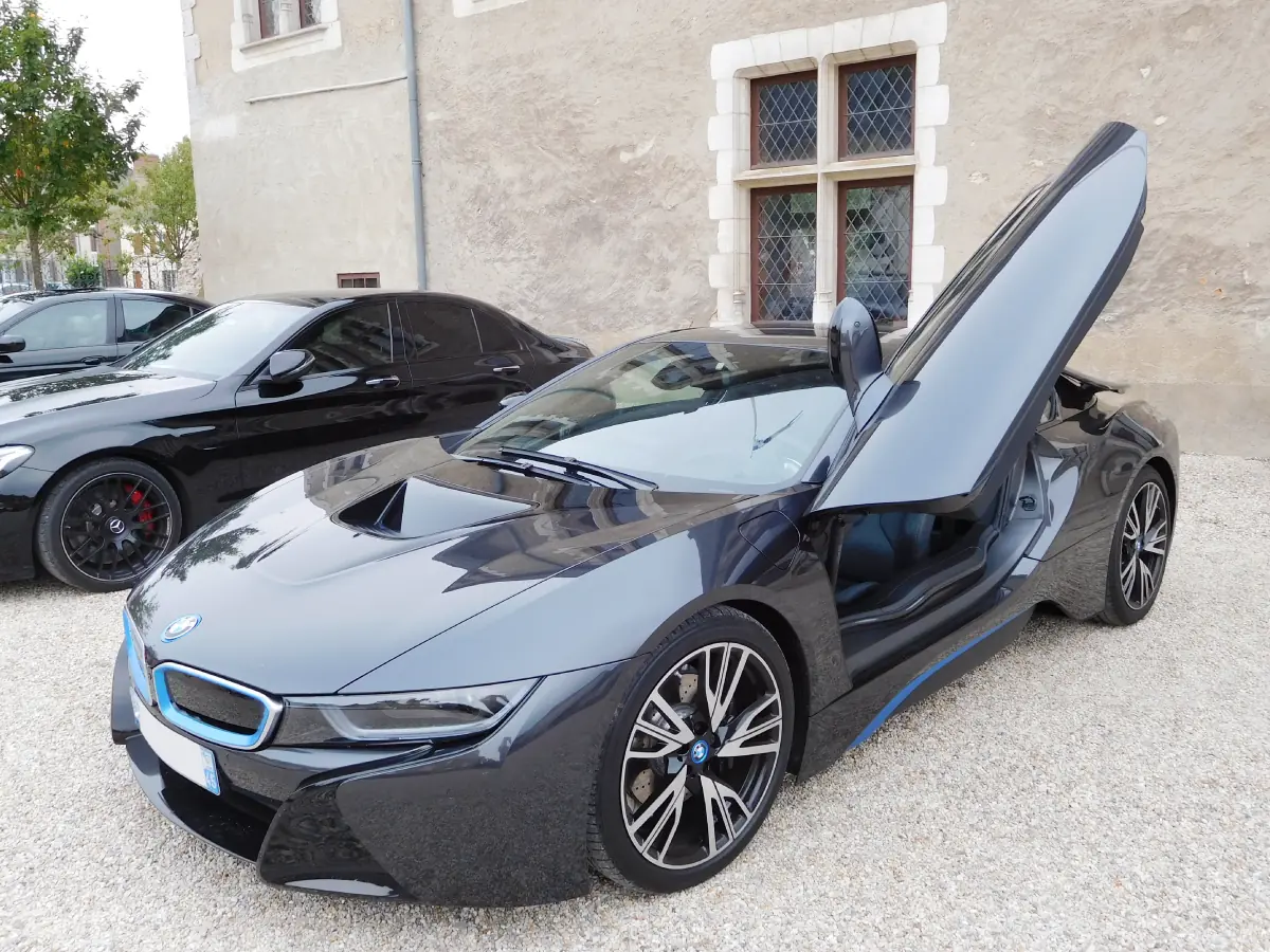 BMW i8 (2014) na cor preta com pequenos detalhes em azul, estacionando em pátio externo com abertura elevada da porta direita.