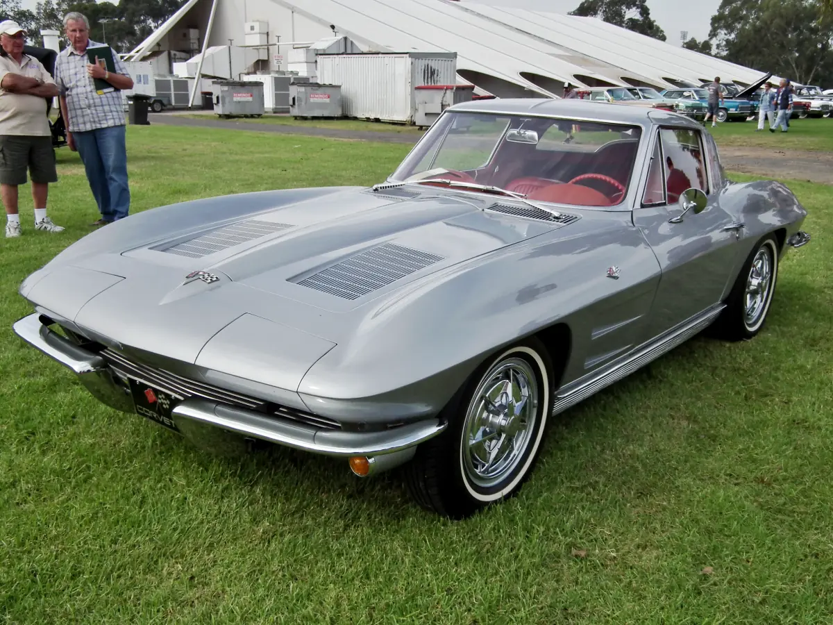 Chevrolet Corvette Stingray (1963) na cor cinza, estacionado sobre gramado, com pessoas na lateral direita do carro apreciando o modelo.