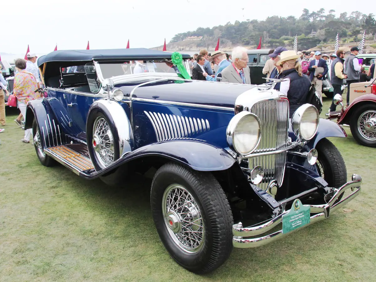 Duesenberg Model J (1928) na cor azul escuro, estacionado sobre gramado, com pessoas circulando e algumas admirando o carro.