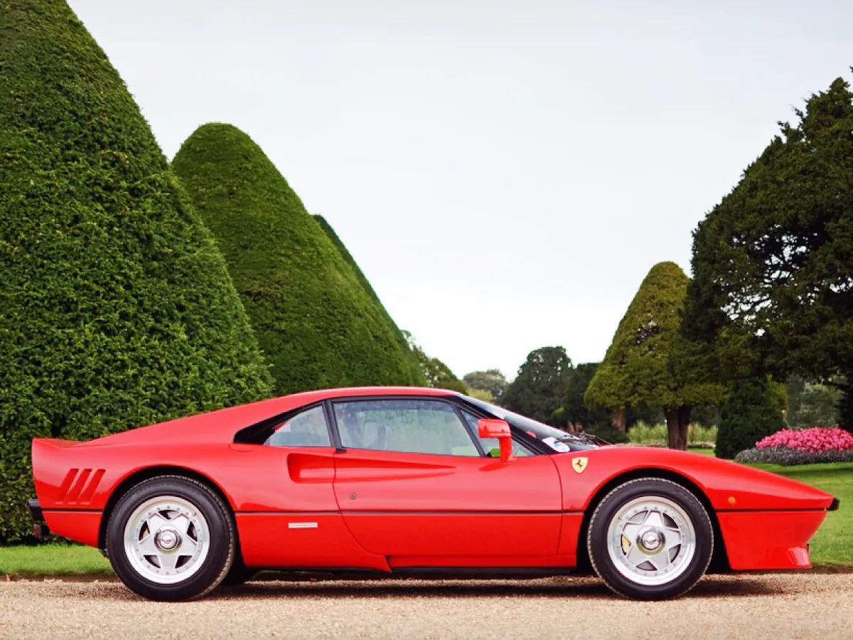 Ferrari 288 GTO (1984) na cor vermelha, estacionado em área verde com vários pinheiros ao fundo.