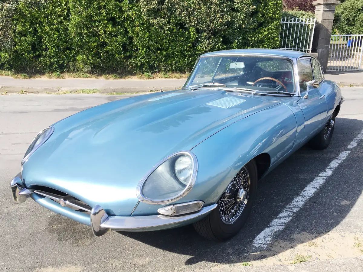 Jaguar E-Type (1961) na cor azul claro, estacionado em ambiente externo, com portões e área verde ao fundo.