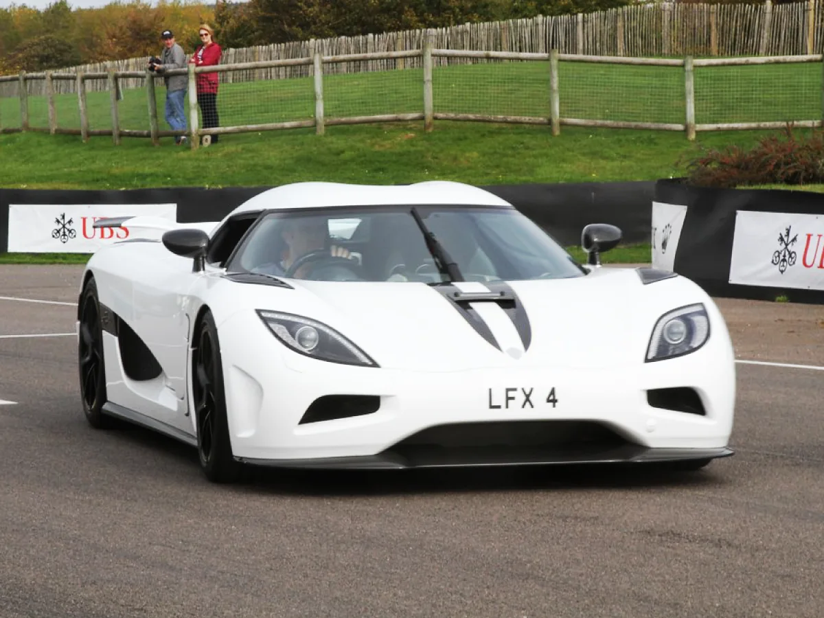 Koenigsegg Agera (2011) na cor branca em pista, com duas pessoas ao fundo admirando o desempenho do carro.