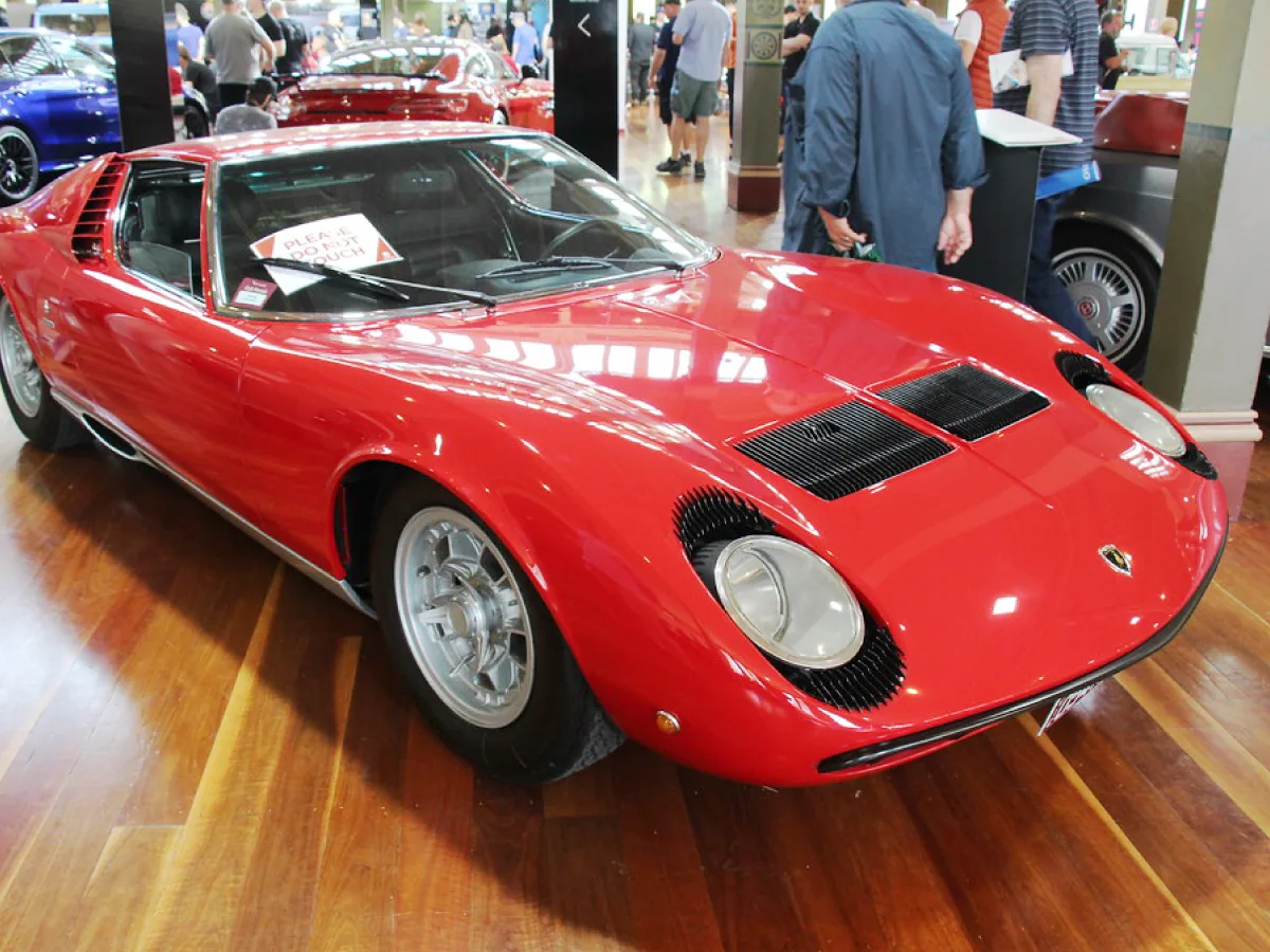 Lamborghini Miura (1966) na cor vermelha, estacionado sobre piso de madeira, com outros carros em exposição e pessoas admirando.