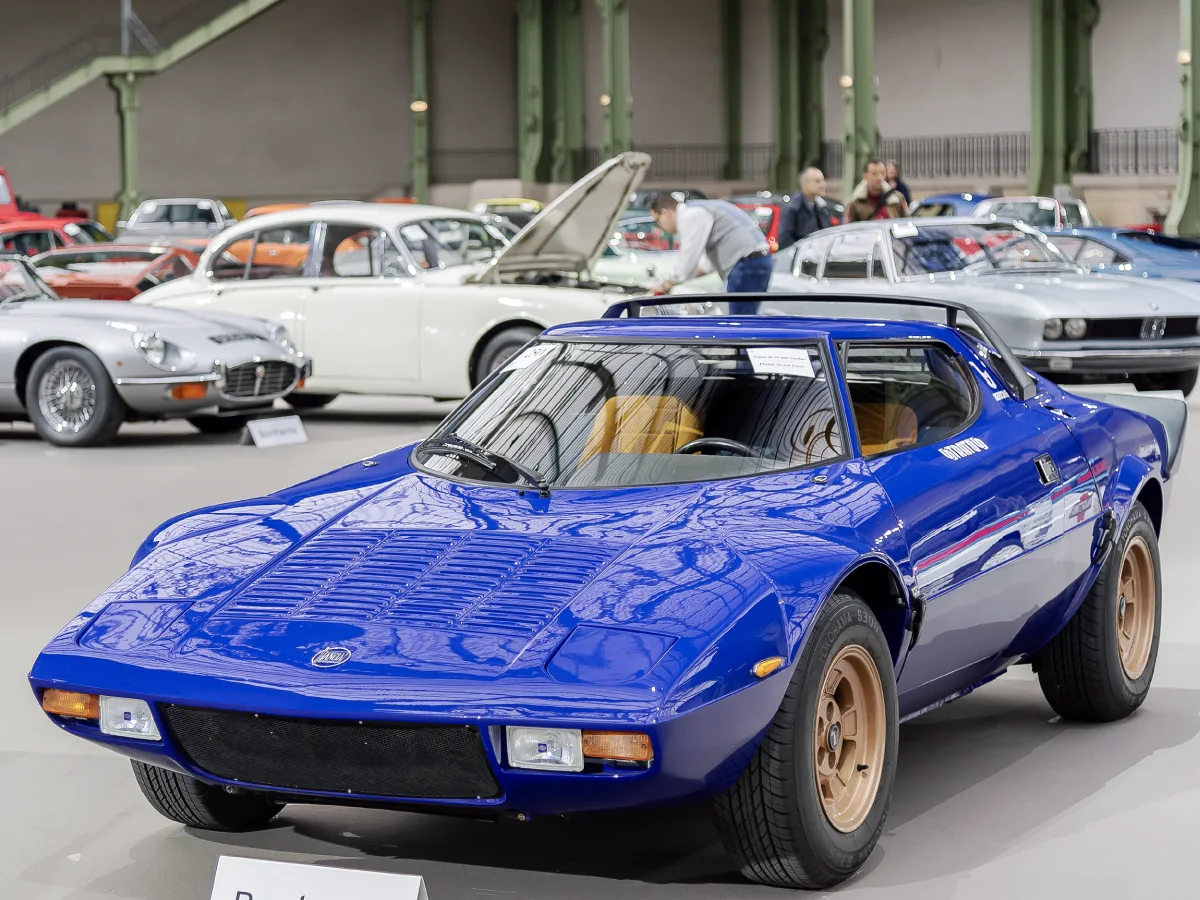 Lancia Stratos (1973) na cor azul royal, estacionado junto a outros carros icônicos.