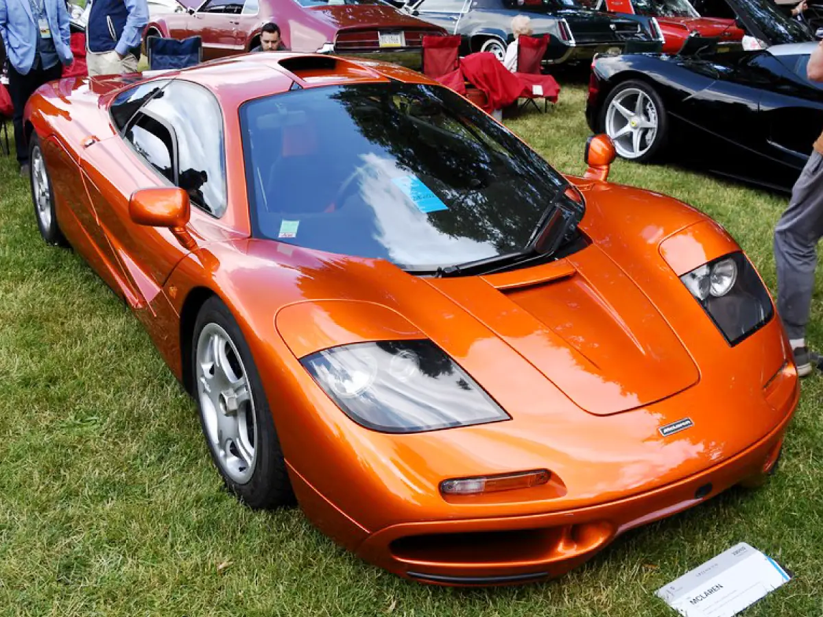 McLaren F1 (1992) na cor laranja, estacionado sobre gramado, tendo outros carros no entorno e pessoas admirando o veículo.