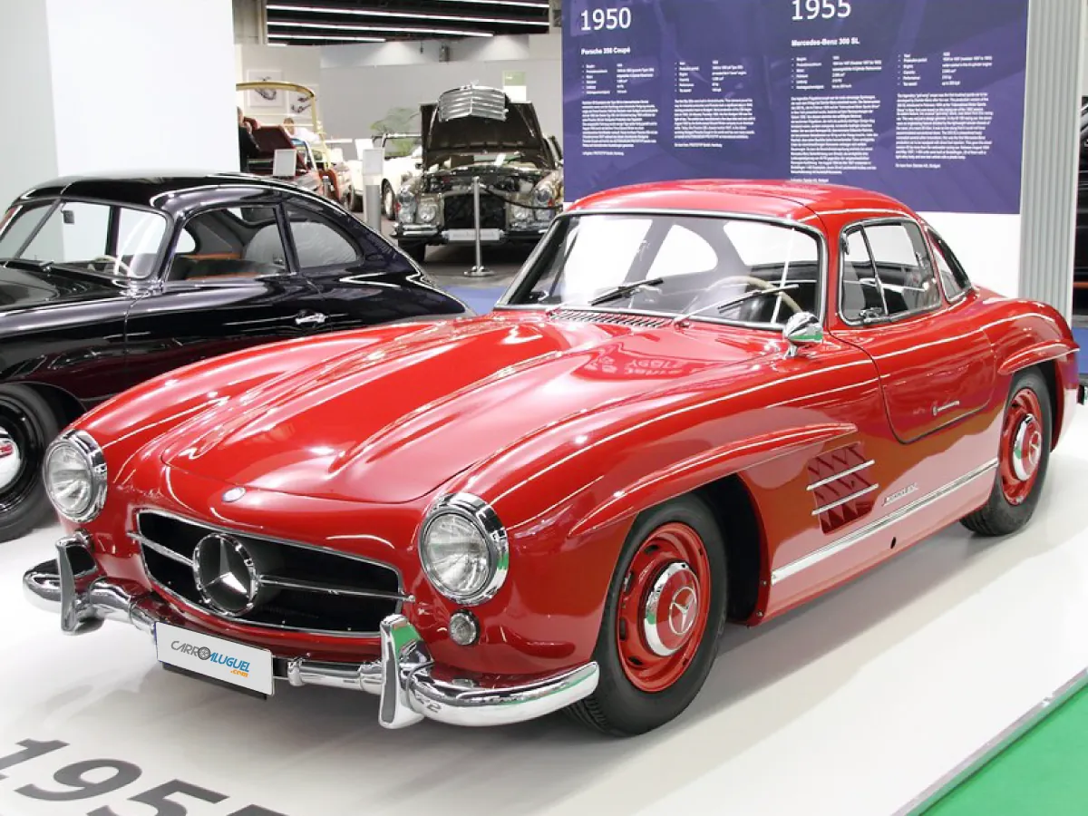 Mercedes-Benz 300SL Gullwing (1954) na cor vermelha, estacionado em feira de exposição, tendo outro carro na cor preta do seu lado direito.