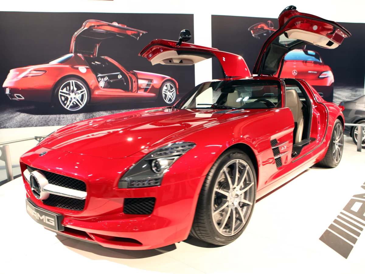 Mercedes-Benz SLS AMG (2010) na cor vermelha, com as portas abertas elevadas e ao fundo um painel fotográfico apresentando o mesmo modelo.