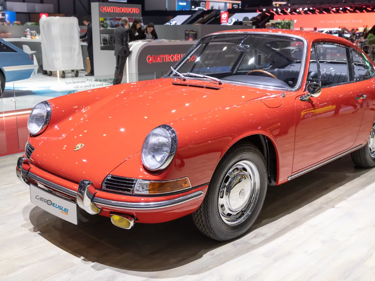 Porsche 911 (1964) na cor vermelha, estacionado em feira de automóvel, com profissionais trabalhando ao fundo.