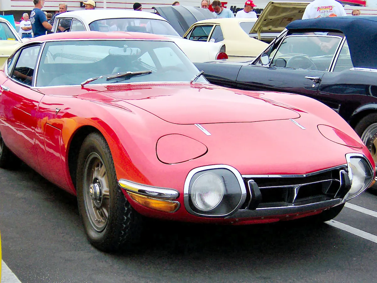 Toyota 2000GT (1967) na cor vermelha estacionado em pátido, tendo ao seu lado outro carro na cor preta com capô conversível.