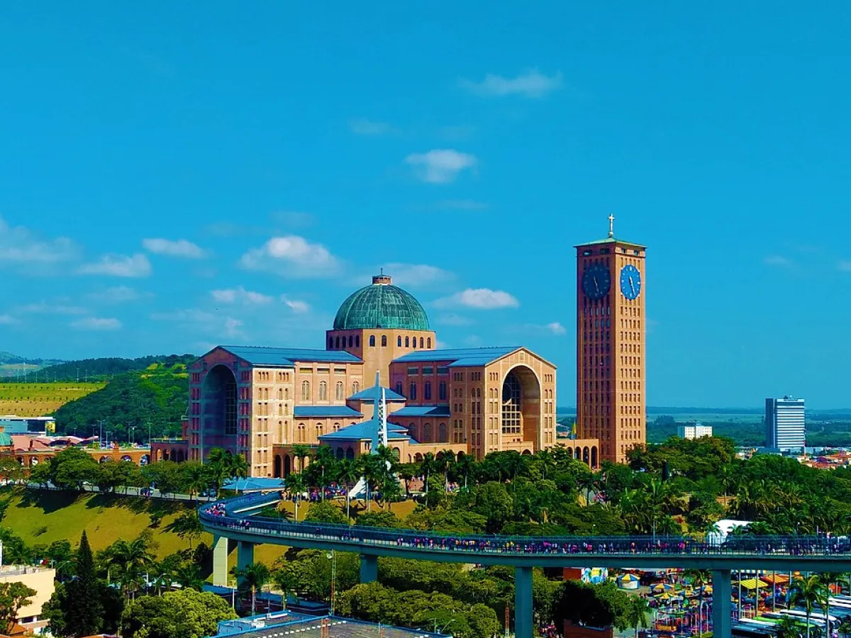 Em destaque segue a Catedral Basícila de Nossa Senhora Aparecida, com ponte repleta de pessoas. Sob a ponte, ruas, espaço arborizado, barracas e automóveis.