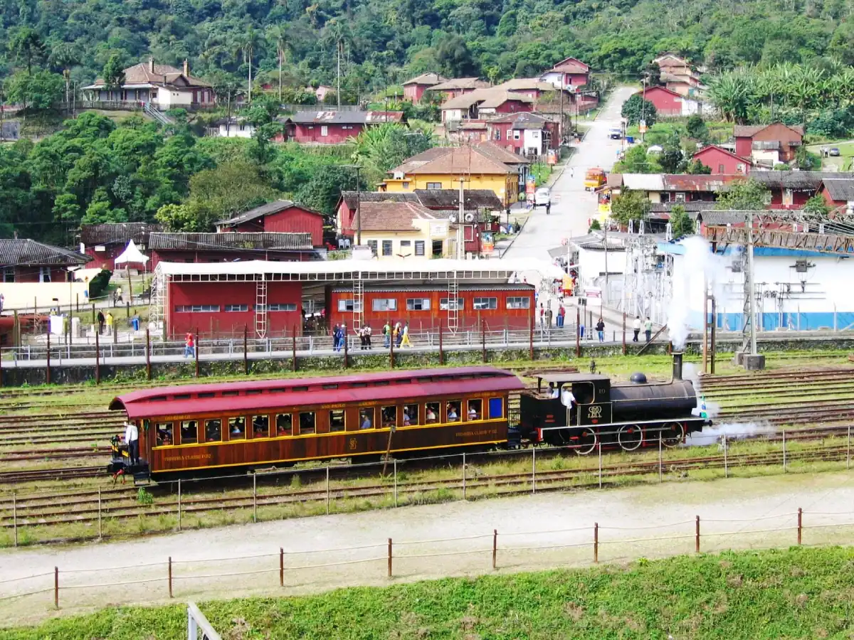 Trem Maria-fumaça sobre trilhos, atração turística na Vila de Paranapiacaba, com passageiros num único vagão e maquinista na locomotiva. Ao fundo há pessoas, casas e carros em uma parte do vilarejo.