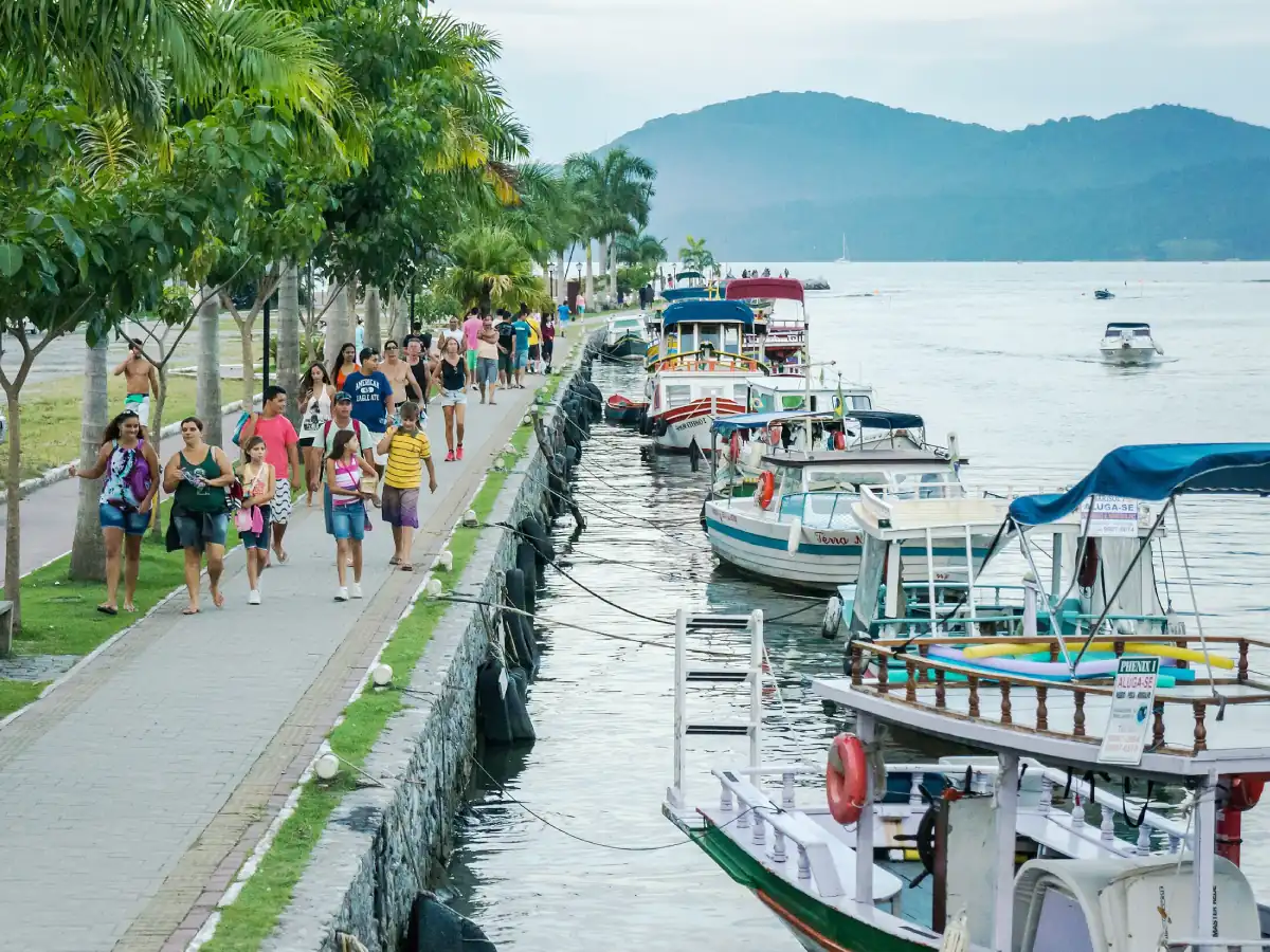 Orla concretada em Paraty com pessoas caminhando sobre a via arborizada, enquanto barcos seguem ancorados em águas calmas da baía. Há um único em movimento.