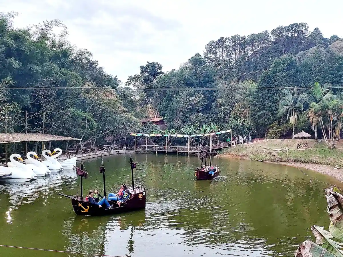 Pedalinhos, alguns estacionados em formato de cisnes, outros sugerindo caravelas carregando turistas, flutuam sobre lago em área temática com muita vegetação e até uma passarela ao fundo.