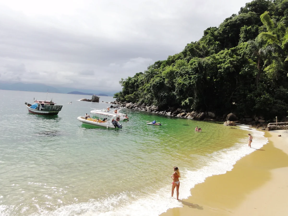 Pequena orla em praia calma de Ubatuba, com turistas entrando na água, embarcações e muita vegetação ao fundo.