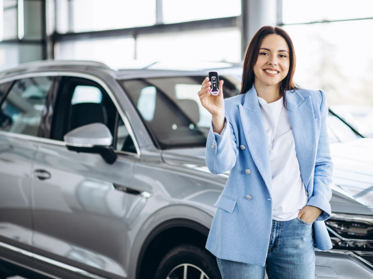 Mulher sorridente motra chave que está na sua mão direita, enquanto a outra mão segue apoiada no bolso. Logo atrás da pessoa há um carro de 4 portas na cor prata.