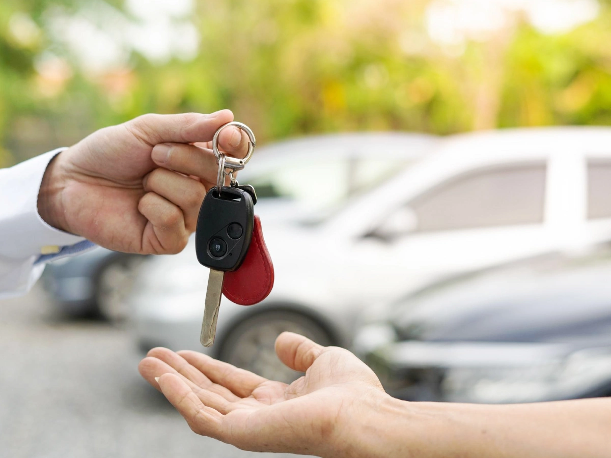 Mão que segura uma chave de automóvel entregando a outra em espera com a palma para cima. Desfocados ao fundo, há alguns veículos.