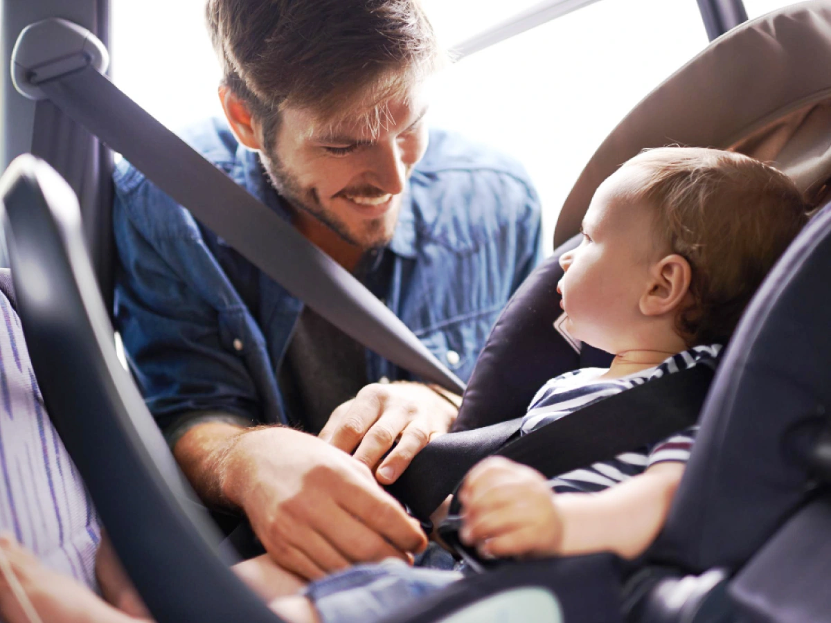 Pai sorridente ajeita o cinto de segurança na cadeirinha de bebê, enquanto observa o filho que segue atento ao movimento.