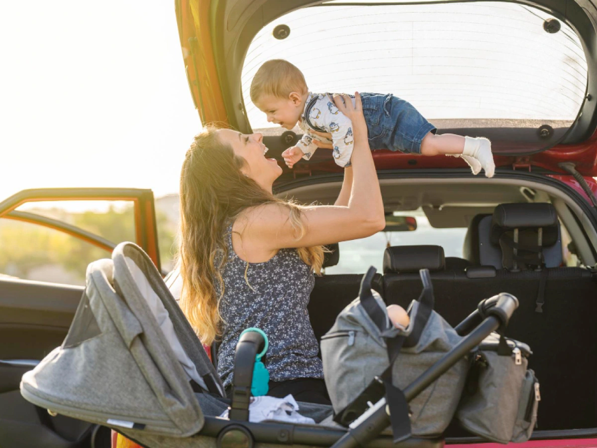 Mãe segura filho nos braços, ambos sorridentes, ele logo após deixar o carrinho de bebê, preparando-se para viagem no carro logo a frente.