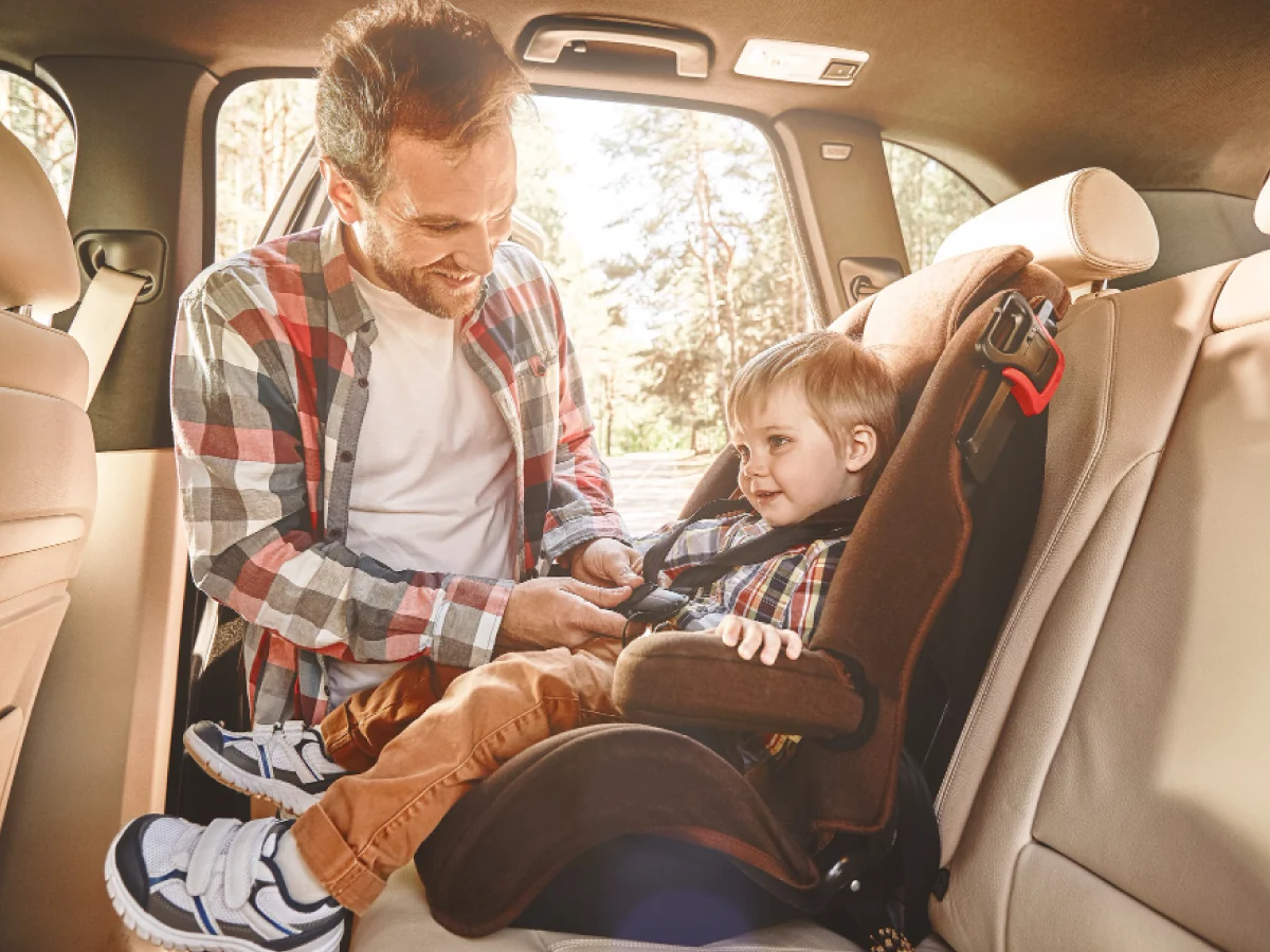 Pai ajeita o cinto de segurança no filho que segue sentado na cadeirinha de bebê, encaixada no banco traseiro de um veículo.