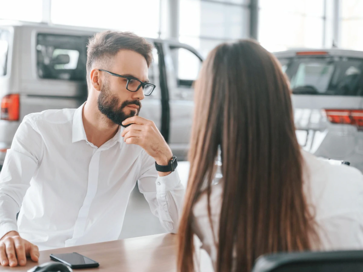 Funcionária conversando com cliente que tem celulcar sobre a mesa e, ao fundo, dois carros estacionados.