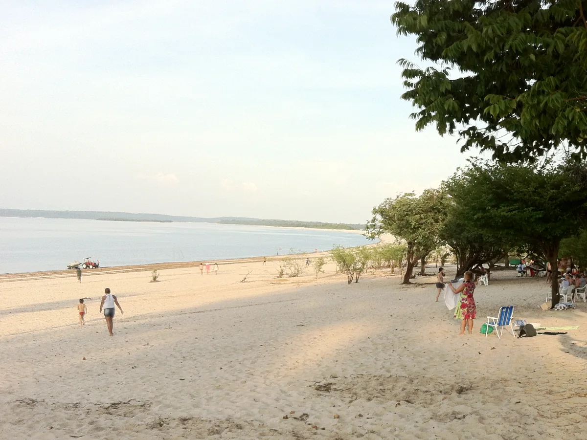 Adultos e crianças passeiam pela orla da Praia do Açutuba, local de vegetação exuberante, areia branca e água escura do Rio Negro.