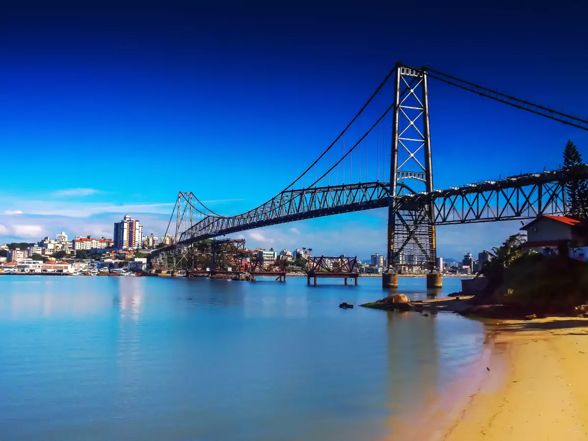 Ponte pênsil Hercílio Luz sobre o Canal do Estreito em Florianópolis. Ao fundo, alguns prédios e o céu azul constrastando com pequena faixa de areia situada no lado da ilha.