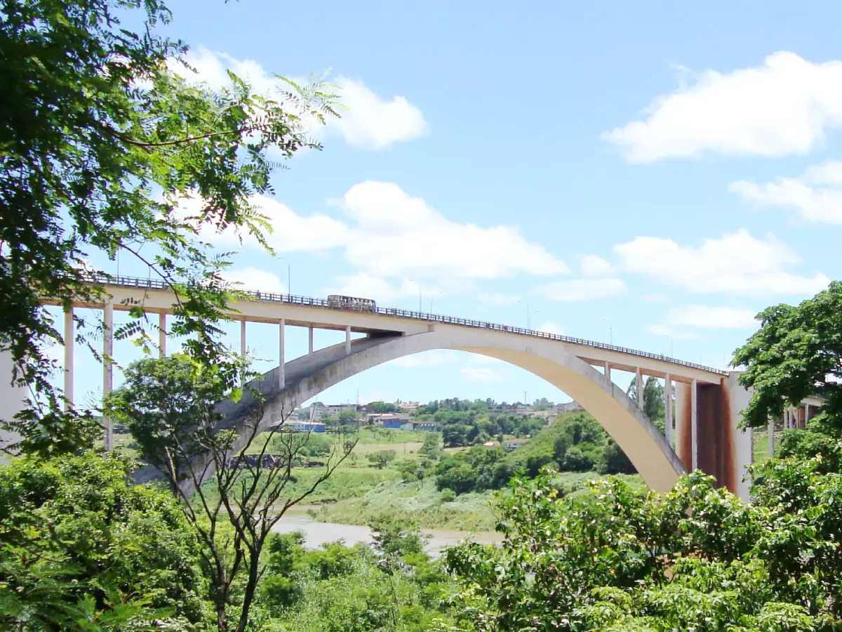 Ponte Internacional da Amizade em meio a uma imensa vegetação e céu azul com nuvens. Há um ônibus atravessando a estrutura de 552 metros de extensão.