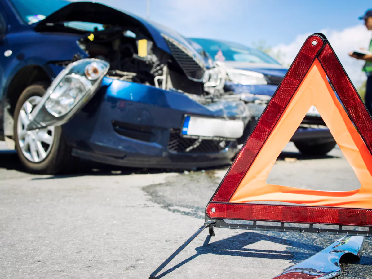 A imagem mostra um triângulo vermelho em destaque, sinalizando o local de um acidente. Ao fundo, um carro azul dafinicado e um segundo carro, ocultado pelo triângulo. Um agente de trânsito aparece parcialmente.