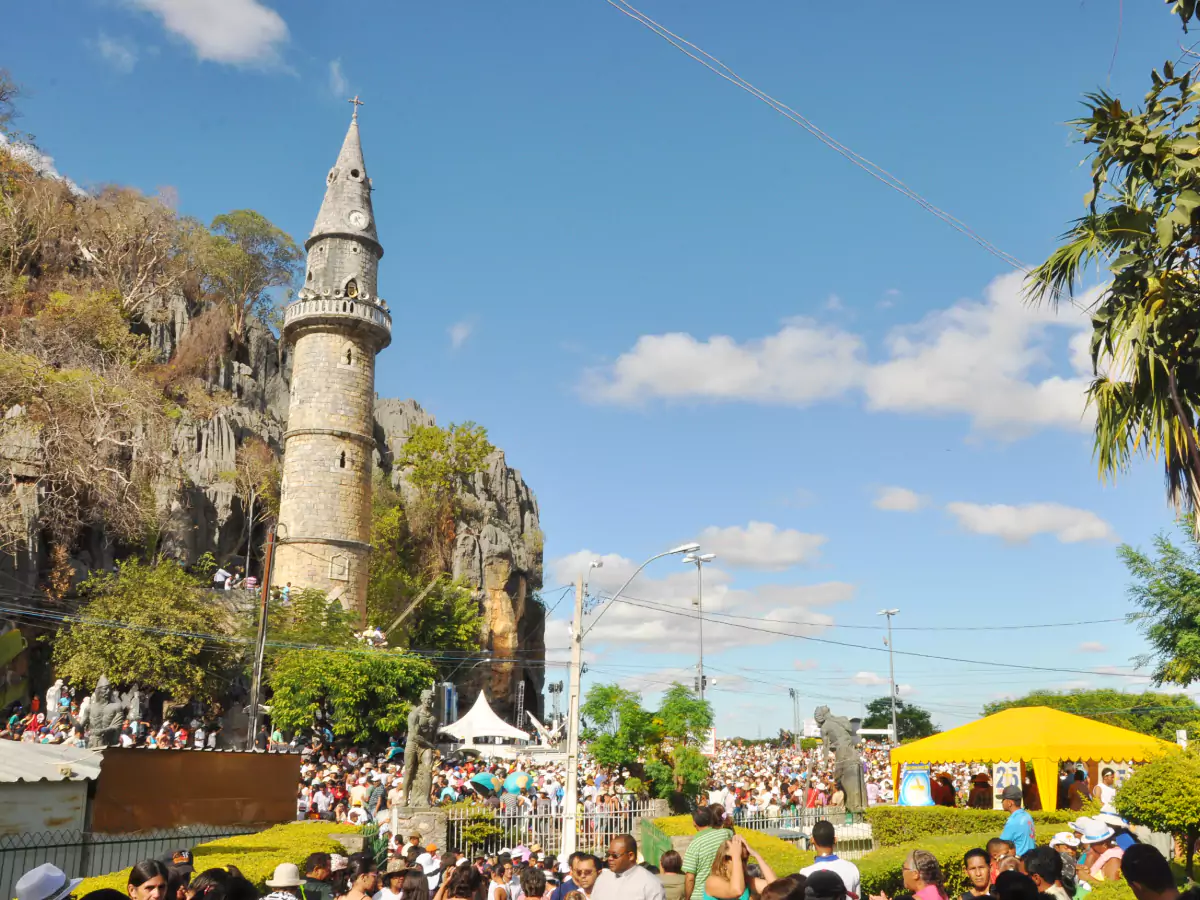 Pessoas circulam por área bastante movimentada na cidade de Bom Jesus da Lapa. Em meio à multidão, há tendas espalhadas. Destaque para a torre do santuário situada bem ao lado de um paredão natural com vegetação variada. O céu encontra-se azul e com poucas nuvens.