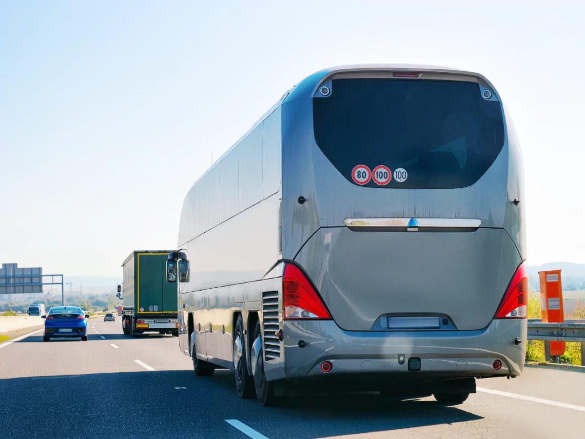 A imagem mostra uma rodovia na qual transitam um ônibus, um caminhão e um carro. O ônibus é cinza e está em primeiro plano. Ao fundo, o céu está azul-claro.