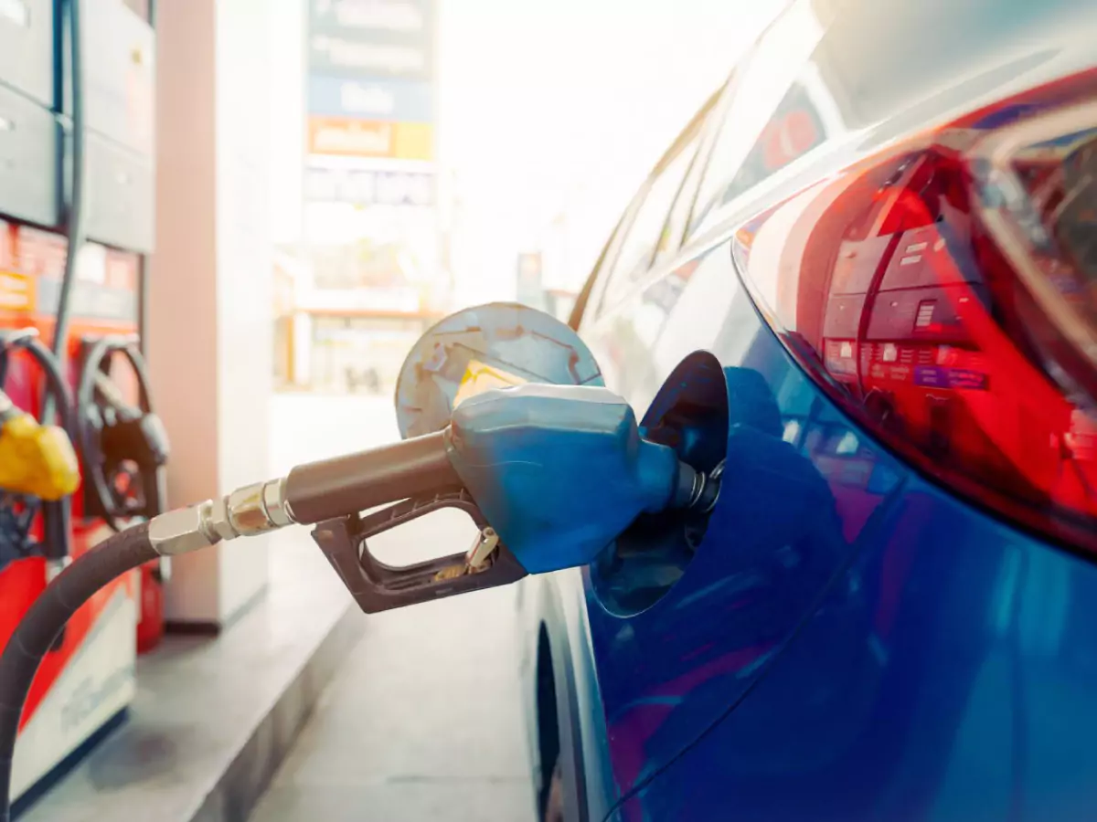 A imagem mostra um carro azul com o tanque de combustível aberto e uma bomba de abastecimento, também azul, em destaque. Ao fundo um posto de abastecimento em vermelho tem bombas amarela e preta. Desfocada ao fundo, uma placa de anúncio de preços dos comburstíveis.