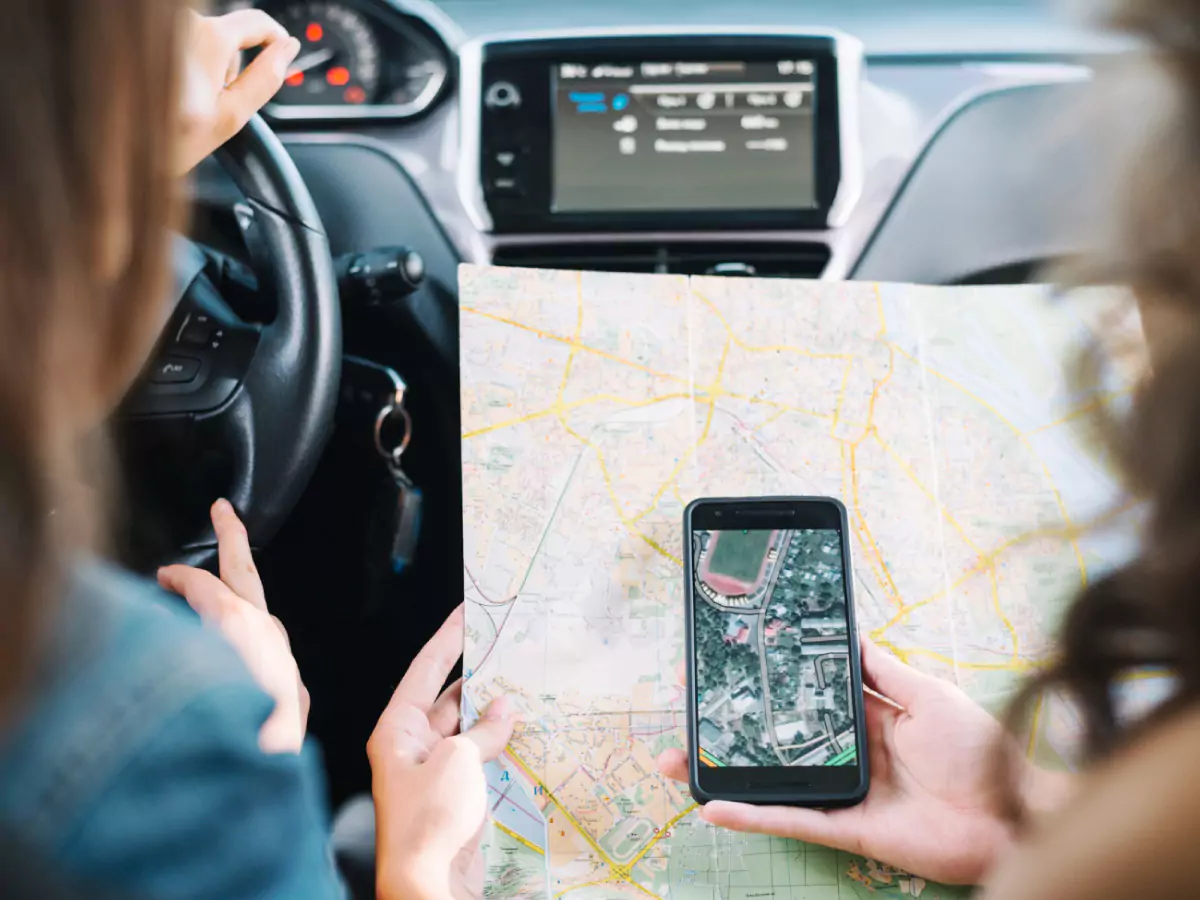 Duas mulheres, ambas dentro de um carro, observam a imagem em destaque no celular, comparando-a com um mapa que segue na mão da passageira.