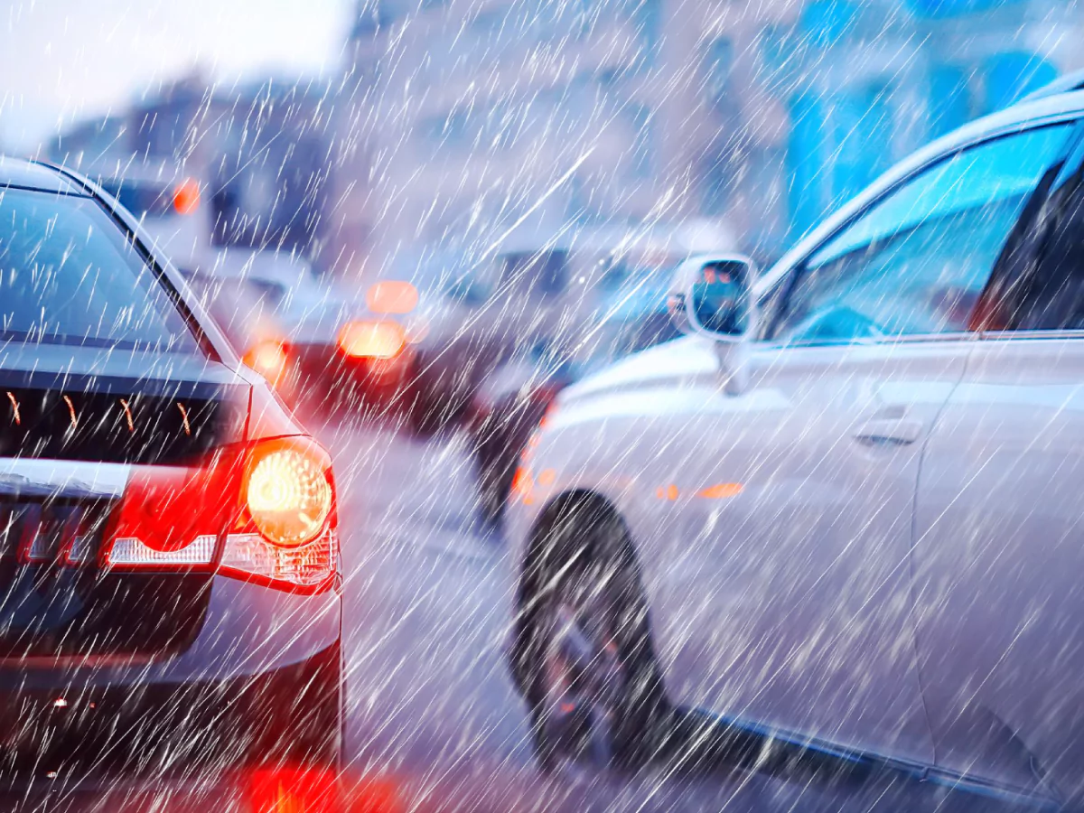 A imagem mostra uma rodovia congestionada durante uma chuva. Em destaque está um carro preto com a luz vermelha de alerta ligada. Ao lado há um carro branco. No fundo, desfocados, estão mais carros com as luzes ligadas e prédios, sugerindo uma via urbana.