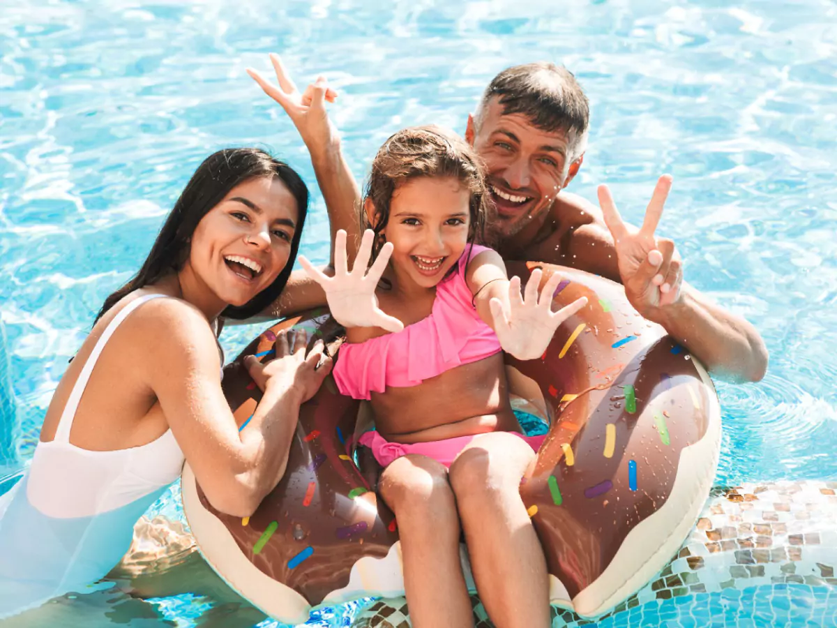 Homem, mulher e criança brincam em água de piscina. Ele está sem camisa, a moça com um colan branco e a criança veste uma roupa cor de rosa e segue sobre uma boia de plástico.