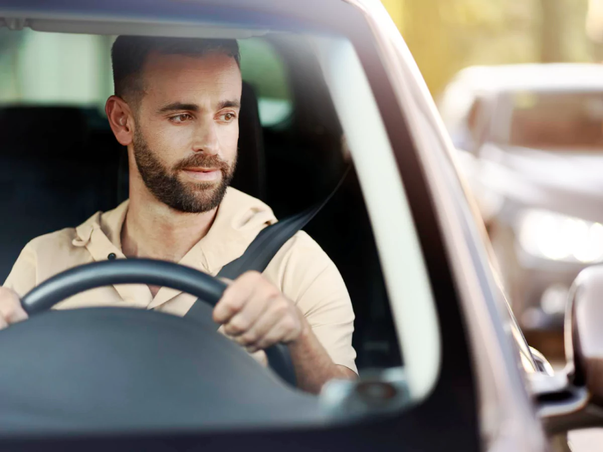 A imagem mostra um homem, de cabelo e barba curtos, camisa marrom-claro, no banco do motorista de um carro. Ele usa o cinto de segurança e tem as duas mãos no volante. Ele olha para o espelho retrovisor. Ao fundo, um veículo desfocado.