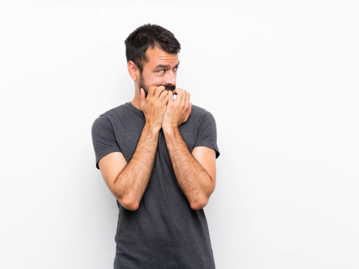 Um homem de cabelo curto e barba, usando uma camisa cinza, olha para a esquerda enquanto rói as unhas. Ele está em um fundo completamente branco.