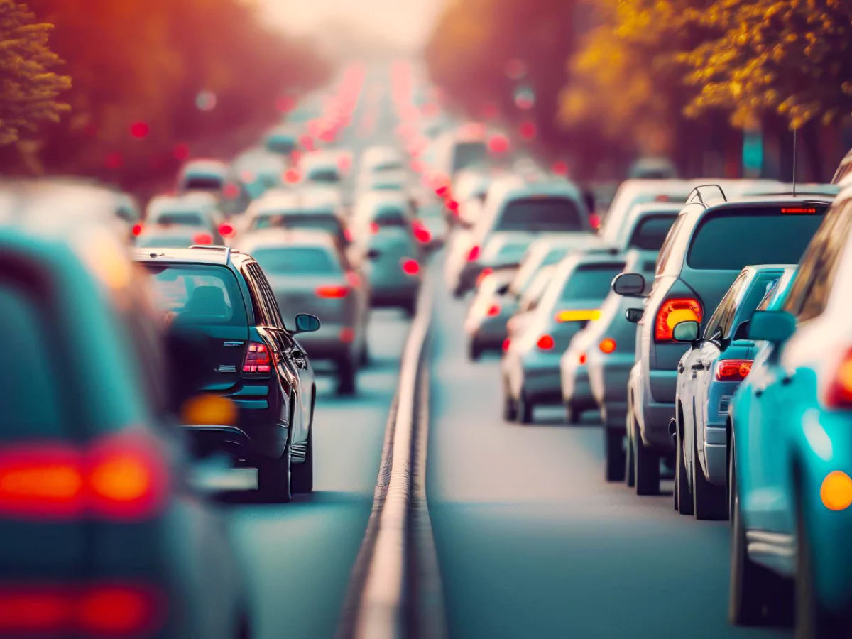 A imagem mostra uma via totalmente ocupada por carros. A fila se estende até o horizonte da imagem, com faróis vermelhos formando uma linha. Nas margens, vegetação no campo superior sugere a presença de árvores.