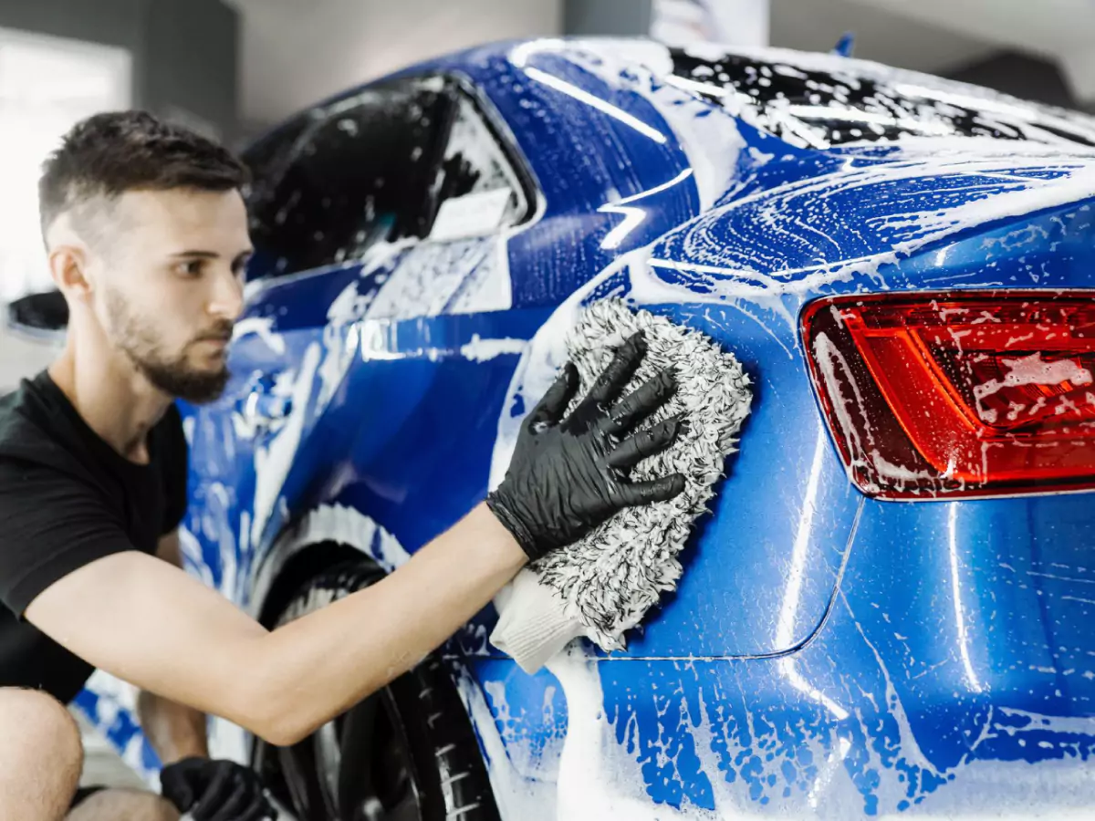 Um homem de cabelo curto e barba, vestido de preto e utilizando luvas pretas, esfrega uma esponja cinza em um carro azul. O carro está com as janelas fechadas, coberto de espuma.